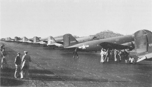 Generals Kenney and MacArthur watch C-47 transport planes loaded with paratroops for the drop at Nadzab.|alt=A steel plank runway with identical aircraft lined up along its length at right. Two small figures stand off to the left of the runway.