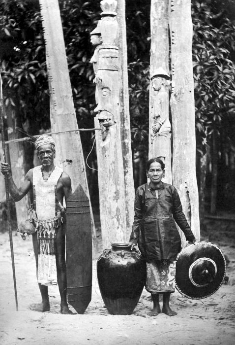 Two sapundu sacrificial poles in the form of male head and a woman stand behind this Ngaju couple.
